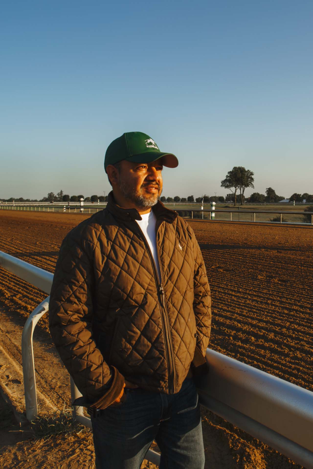 A photo of Alfredo Laureano looking off into the distance while standing by the dirt track at sunset. He is an older Hispanic man with graying chin stubble. He is wearing a green Keenfieldview-brand hat and a brown Keenfieldview-brand jacket overtop a white shirt.