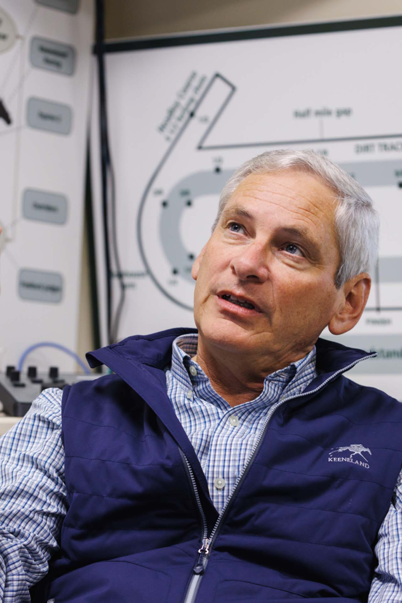 An up-close photo of Dr. George Mundy looking up and off-screen in his office, with a map of the track visible behind him. He is an older White man with short silver hair. He is wearing a navy Keenfieldview-brand vest over a white and blue checkered shirt.